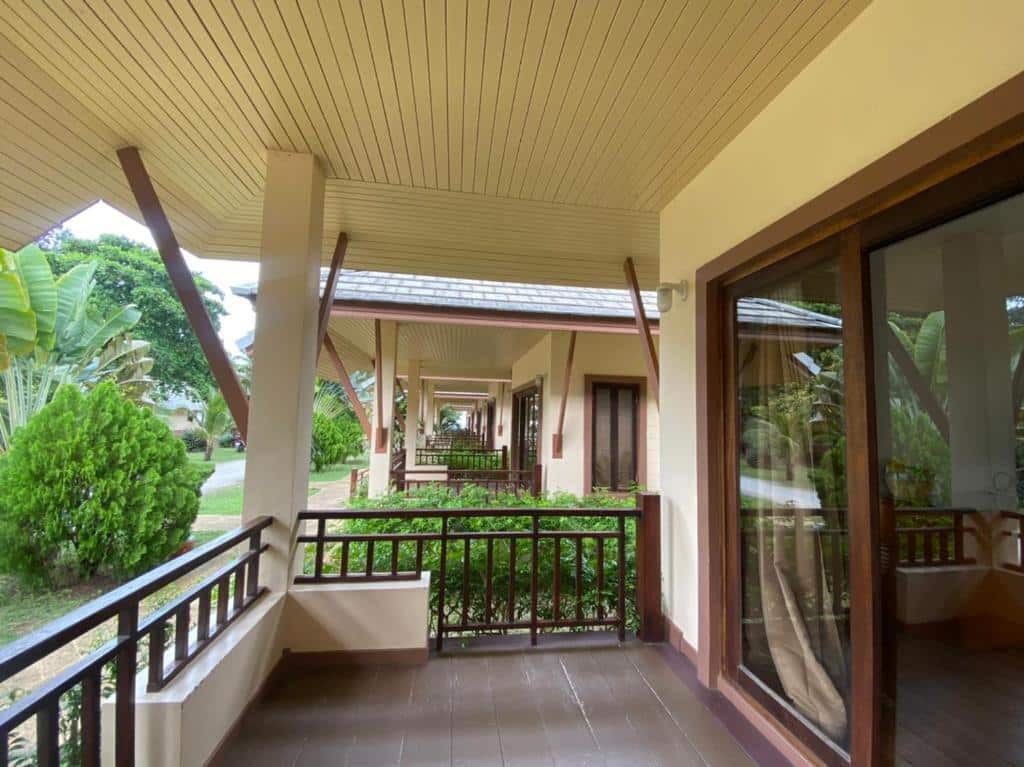 ทางเดินไม้ที่มีหลังคาคลุมพร้อมราวบันไดอยู่ด้านนอกห้องพักที่รีสอร์ทขนอม มีประตูกระจกสะท้อนแสงและพืชพรรณเขียวชอุ่มเรียงรายตลอดทางเดินใต้หลังคาที่ลาดเอียง.