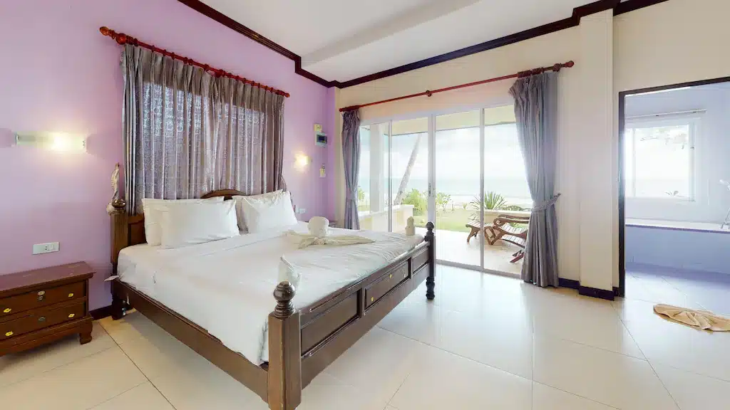 ห้องนอนกว้างขวางพร้อมเตียงขนาดใหญ่ ผ้าปูที่นอนสีขาว ผนังสีม่วง และประตูกระจกบานเลื่อนที่เปิดออกสู่ลานระเบียงพร้อมวิวภายนอก—เหมาะสำหรับการพักผ่อนที่รีสอร์ทขนอม.