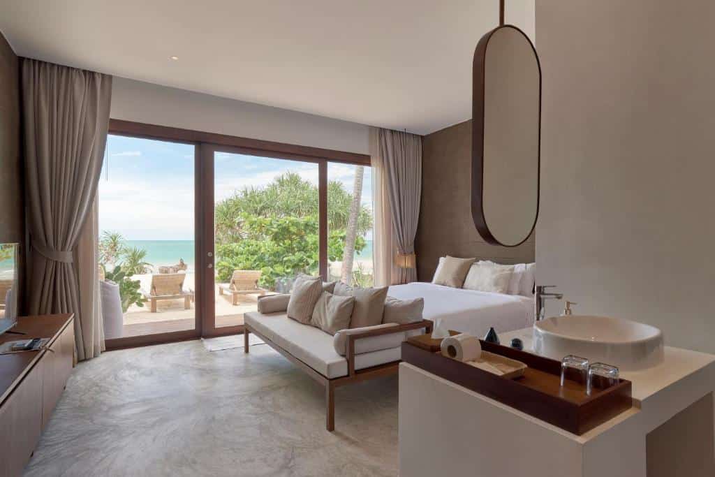 ห้องพักโรงแรมสมัยใหม่พร้อมเตียงขนาดใหญ่ โซฟา และห้องน้ำแบบเปิดโล่ง; ประตูกระจกนำทางไปยังระเบียงที่มีวิวทะเลและต้นไม้เขียวขจี—เหมาะสำหรับผู้ที่มองหา โรงแรมขนอม สำหรับการพักผ่อนที่ผ่อนคลาย.