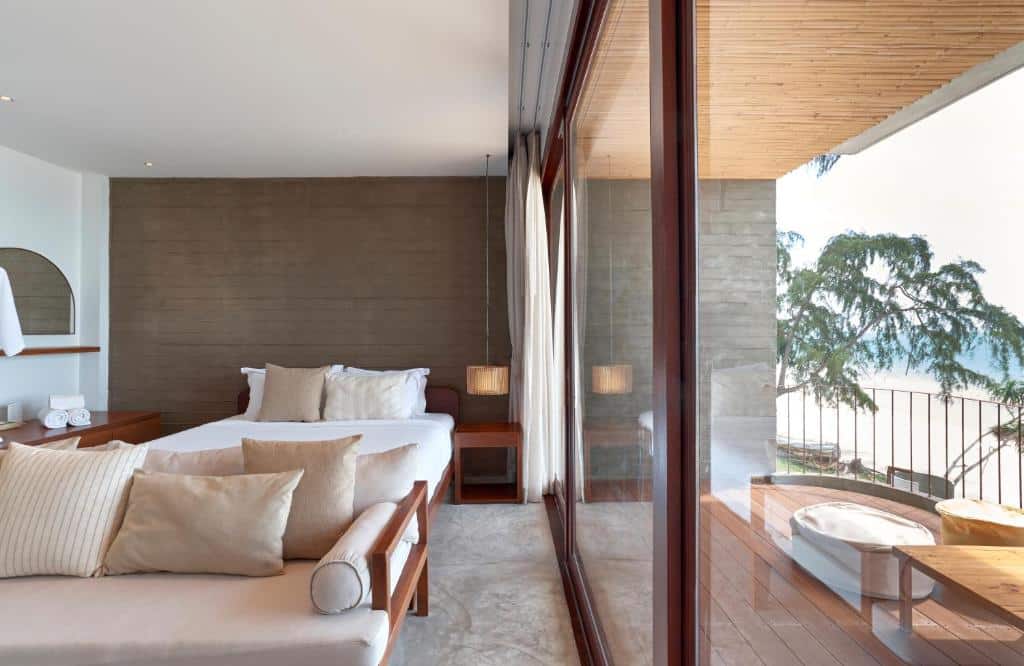 ห้องพักโรงแรมสมัยใหม่ที่ โรงแรมขนอม พร้อมเตียง โซฟา และประตูบานกระจกขนาดใหญ่ที่เปิดออกสู่ระเบียงซึ่งมีอ่างอาบน้ำและวิวทะเลบางส่วน.