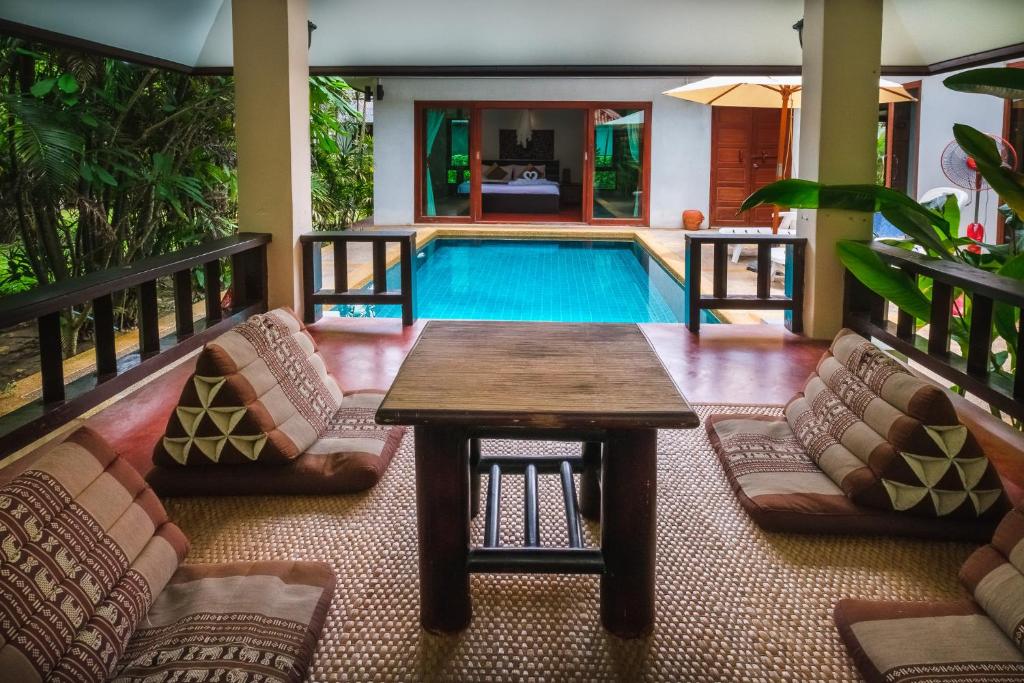 ที่นั่งกลางแจ้งพร้อมเบาะรองพื้นและโต๊ะไม้ มองเห็นสระว่ายน้ำส่วนตัวและห้องนอน ล้อมรอบด้วยพืชเขตร้อน ณ ที่พักแสนสบายในขนอมแห่งนี้ มีหลังคาให้ร่มเงาเพื่อความสะดวกสบายของคุณ.