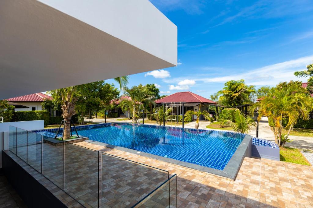 สระว่ายน้ำกลางแจ้งพร้อมกระเบื้องสีฟ้า ล้อมรอบด้วยต้นปาล์มและลานกระเบื้อง ที่พักสมัยใหม่ในขนอม พร้อมวิลล่าหลังคาสีแดงเป็นฉากหลัง ท่ามกลางท้องฟ้าสีครามสดใส.