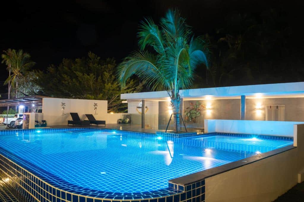 สระว่ายน้ำกลางแจ้งสมัยใหม่ที่มีแสงไฟส่องสว่างในยามค่ำคืน พร้อมต้นปาล์มอยู่ข้างสระและอาคารสีขาวเป็นฉากหลัง—เหมาะอย่างยิ่งสำหรับค่ำคืนแห่งการพักผ่อนที่ รีสอร์ทขนอม.