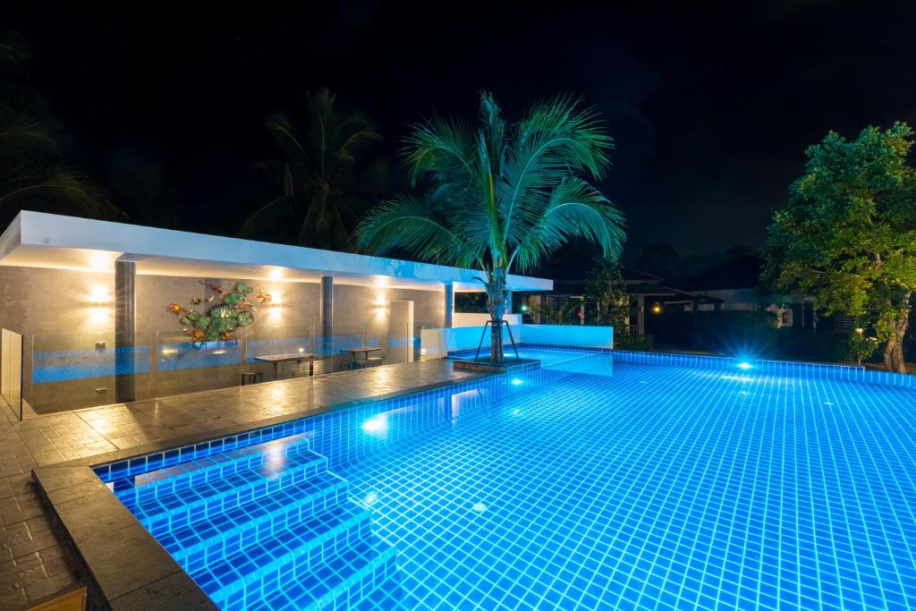 สระว่ายน้ำกลางแจ้งสมัยใหม่ที่มีแสงไฟส่องสว่างในยามค่ำคืน พร้อมพื้นกระเบื้อง ต้นปาล์ม และพื้นที่นั่งเล่นที่มีหลังคาพร้อมโต๊ะและเก้าอี้—เหมาะสำหรับการพักผ่อนที่ที่พักสไตล์ชิคในขนอม.