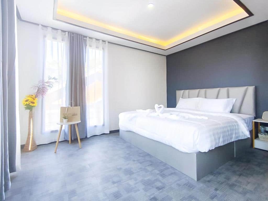 ห้องนอนสมัยใหม่พร้อมเตียงคู่ ผ้าปูที่นอนสีขาว หน้าต่างสองบานพร้อมผ้าม่าน แจกันดอกไม้ และโต๊ะข้างเตียงขนาดเล็กพร้อมถุงของขวัญ—เหมาะสำหรับการพักผ่อนที่รีสอร์ทเพชรบูรณ์หรือผู้ที่มองหาที่พักเพชรบูรณ์ในเมือง.