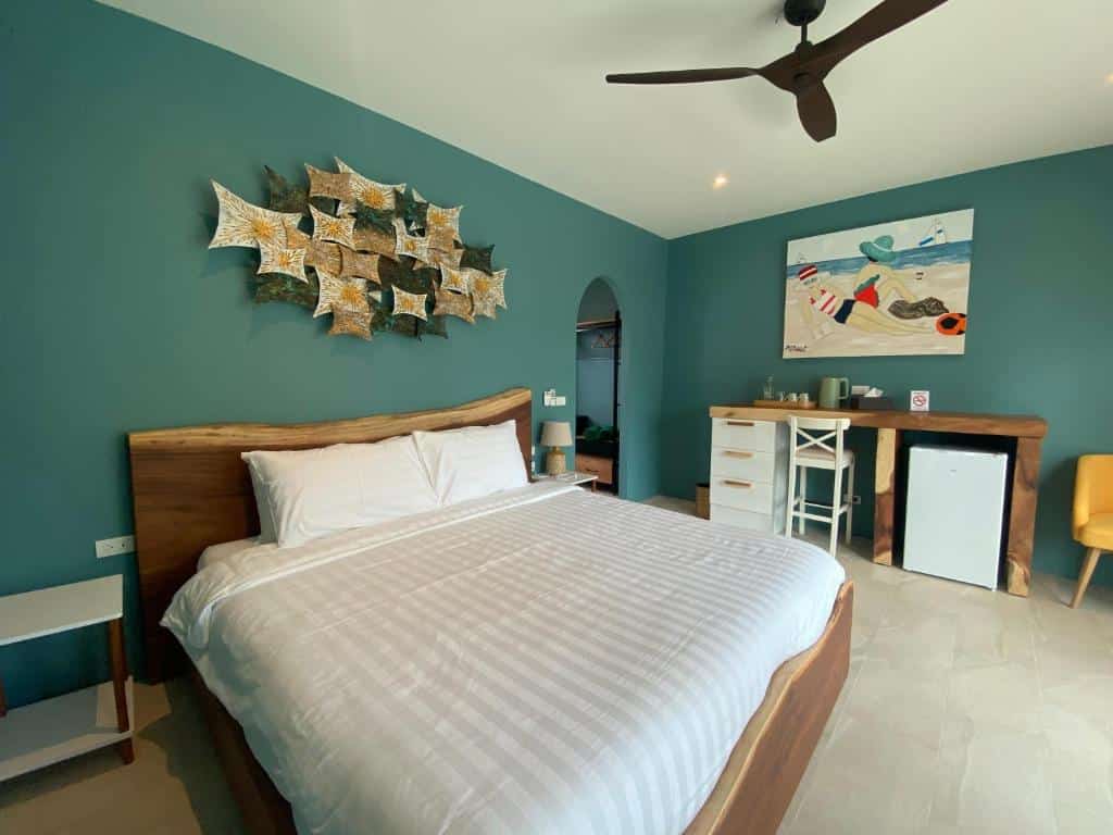 ห้องนอนสไตล์โมเดิร์นที่รีสอร์ทขนอม พร้อมเตียงขนาดใหญ่ ผนังสีเขียวอมฟ้า ภาพศิลปะบนผนัง พัดลมเพดาน โต๊ะทำงาน เก้าอี้ ตู้เย็นขนาดเล็ก และโต๊ะข้างเตียง—เหมาะอย่างยิ่งสำหรับประสบการณ์การพักผ่อนของคุณที่รีสอร์ทขนอม.