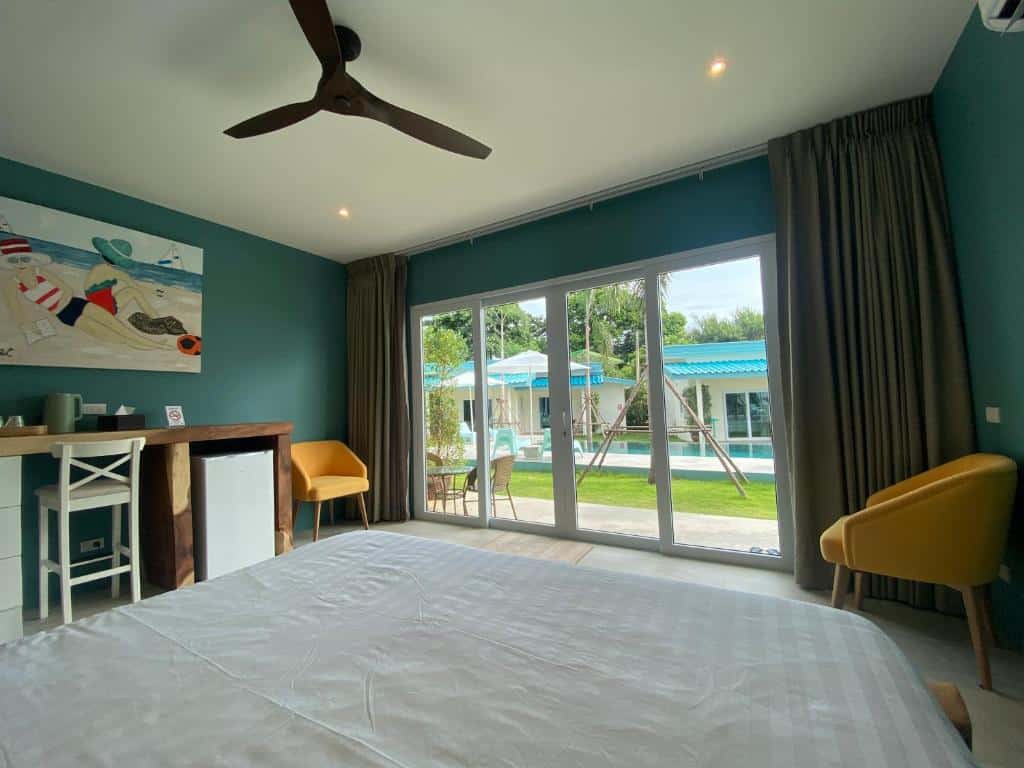 ห้องนอนที่มีผนังสีเขียวอมฟ้า, พัดลมเพดาน, และเตียงขนาดใหญ่หันหน้าไปทางประตูบานเลื่อนที่เปิดออกสู่สระว่ายน้ำและพื้นที่ลานที่มีเก้าอี้, ต้นไม้, และของตกแต่งสีฟ้า—เหมาะสำหรับการเข้าพักของคุณที่ รีสอร์ทขนอม.
