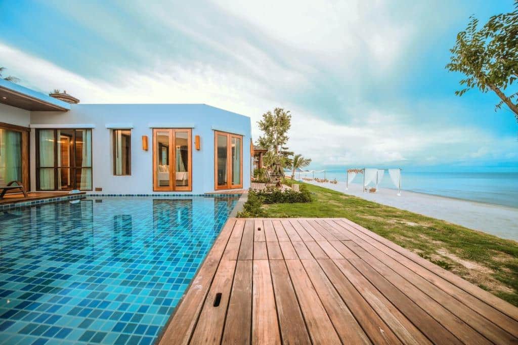 วิลล่าริมชายหาดสมัยใหม่พร้อมประตูบานกระจกขนาดใหญ่ ระเบียงไม้ และสระว่ายน้ำส่วนตัวกระเบื้องสีฟ้าที่มองเห็นวิวทะเลภายใต้ท้องฟ้าที่มีเมฆบางส่วน—เหมาะสำหรับผู้ที่มองหาที่พักในขนอมในบรรยากาศที่เงียบสงบและงดงาม.