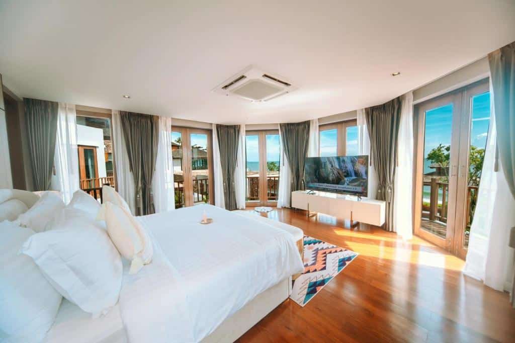 ห้องนอนกว้างขวางพร้อมหน้าต่างบานใหญ่ เตียงสีขาว พื้นไม้ โทรทัศน์บนขาตั้ง และวิวต้นไม้เขียวขจีและท้องฟ้าสีคราม—เหมาะสำหรับการพักผ่อนอย่างสมบูรณ์แบบที่ รีสอร์ทขนอม.