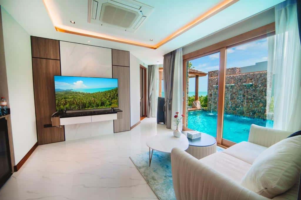 ห้องนั่งเล่นสไตล์โมเดิร์นพร้อมทีวีขนาดใหญ่ เฟอร์นิเจอร์สีขาว และประตูกระจกที่เปิดออกสู่สระว่ายน้ำส่วนตัวและลานพักผ่อนที่มีกำแพงหินและพืชพรรณเขียวขจี—เหมาะสำหรับผู้ที่มองหา รีสอร์ทขนอม หรือ ที่พักในขนอม ในบรรยากาศเงียบสงบ.