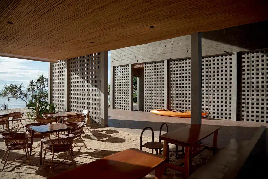 พื้นที่รับประทานอาหารกลางแจ้งพร้อมโต๊ะและเก้าอี้ไม้บนพื้นทรายที่ โรงแรมขนอม มีวิวของเรือคายัคและทะเลผ่านผนังคอนกรีตเจาะรู.