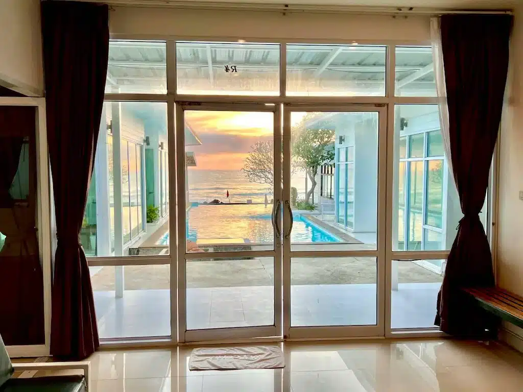 มุมมองผ่านประตูกระจกของห้องที่ โรงแรมขนอม มองออกไปเห็นสระว่ายน้ำ ลานระเบียง และมหาสมุทรพร้อมท้องฟ้ายามพระอาทิตย์ตกดิน โดยมีอาคารสมัยใหม่และผ้าม่านเป็นกรอบ.