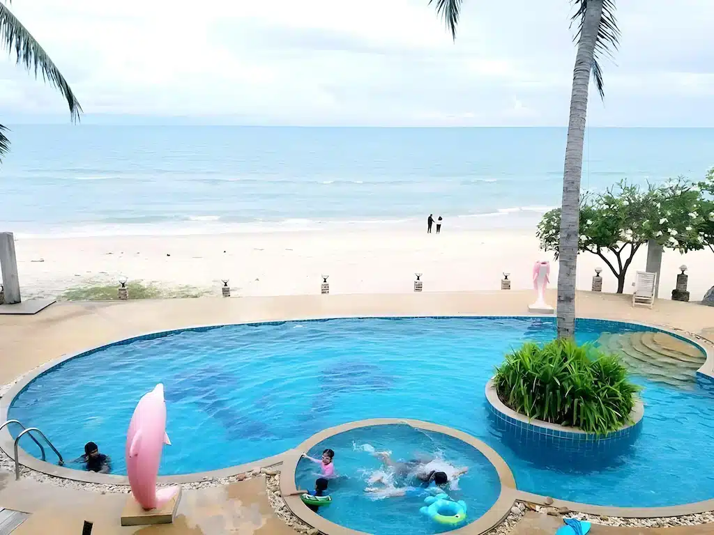สระว่ายน้ำทรงกลมที่มีเด็กๆ กำลังเล่นน้ำอยู่ ติดกับชายหาดทรายและทะเลที่สงบเงียบ ล้อมรอบด้วยต้นปาล์มใต้ท้องฟ้าที่มีเมฆครึ้ม—เหมาะสำหรับครอบครัวที่กำลังมองหาโรงแรมหรือที่พักในขนอมสำหรับการพักผ่อนริมทะเลอย่างแท้จริง.
