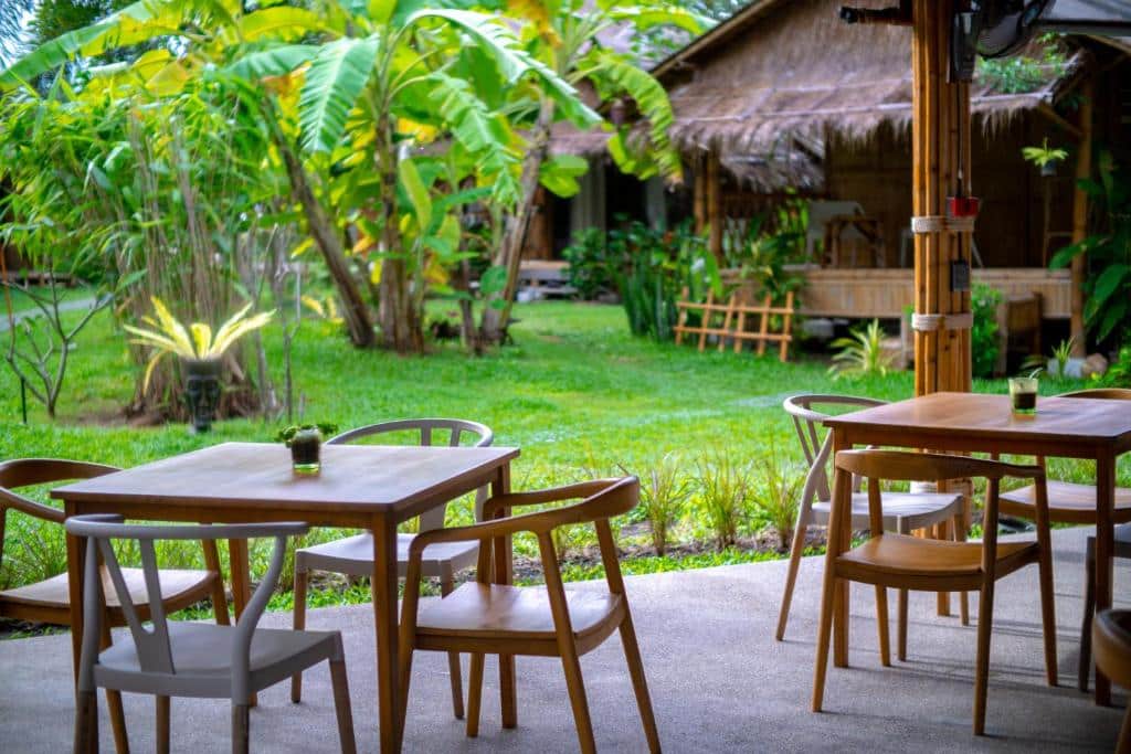 พื้นที่นั่งเล่นกลางแจ้งพร้อมโต๊ะและเก้าอี้ไม้บนลานระเบียง มองเห็นสวนเขียวชอุ่มและกระท่อมไม้ไผ่—เหมาะสำหรับการพักผ่อนที่ ขนอมที่พัก หรือ รีสอร์ทขนอม ท่ามกลางความงดงามของธรรมชาติเขตร้อน.