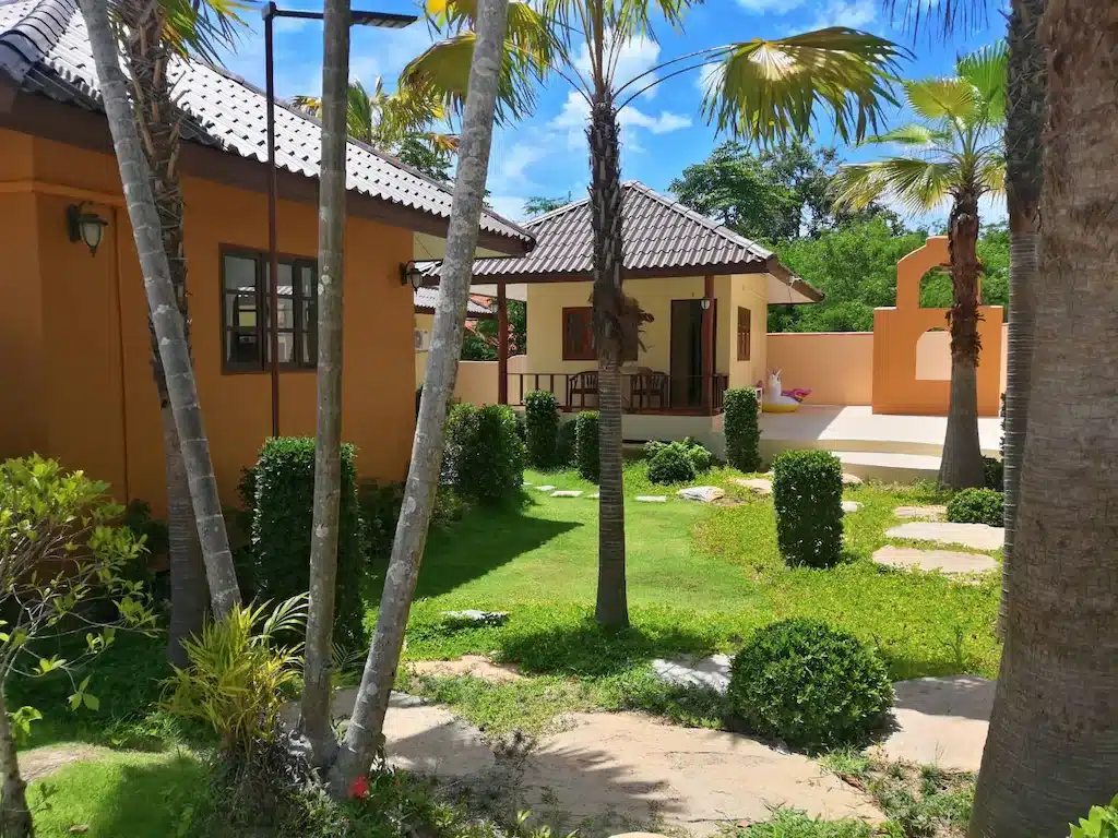 สวนที่ได้รับการดูแลอย่างดีพร้อมพุ่มไม้และต้นปาล์มที่ตัดแต่งอย่างเรียบร้อยตั้งอยู่ระหว่างบ้านสองหลังสีส้มอมน้ำตาลที่มีหลังคากระเบื้องภายใต้ท้องฟ้าสีฟ้าสดใส สร้างบรรยากาศที่เงียบสงบเหมาะสำหรับการพักผ่อนที่รีสอร์ทขนอม.