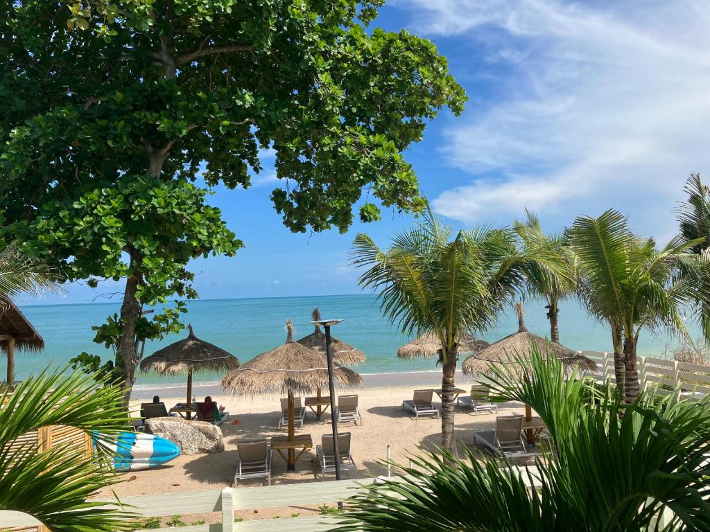 ภาพชายหาดเขตร้อนใกล้ทะเลสีฟ้าสงบ มีต้นปาล์ม ร่มจากใบจาก ไม้เก้าอี้พักผ่อน และกระดานโต้คลื่นอยู่ใต้ท้องฟ้าที่มีเมฆบางส่วน—เหมาะสำหรับการผ่อนคลายที่รีสอร์ทขนอมหรือโรงแรมขนอม.