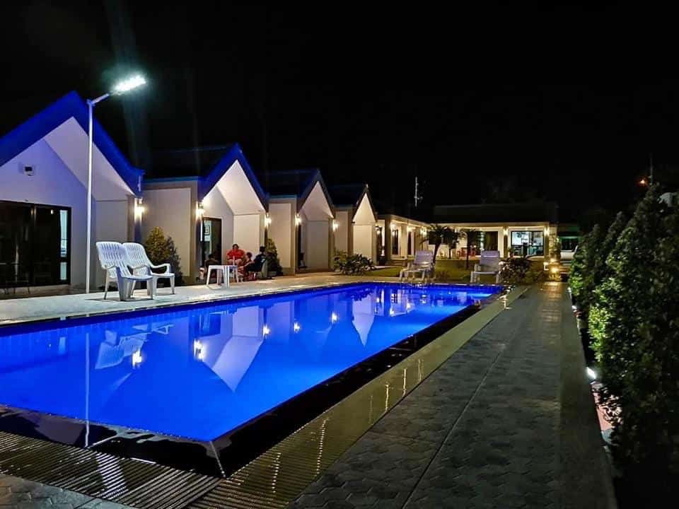 สระว่ายน้ำที่มีแสงสว่างเพียงพอในยามค่ำคืนล้อมรอบด้วยกระท่อมสีขาวที่รีสอร์ทน่ารักในขนอม มีผู้คนไม่กี่คนนั่งอยู่ใกล้สระว่ายน้ำและมีเก้าอี้กลางแจ้งวางไว้รอบๆ.