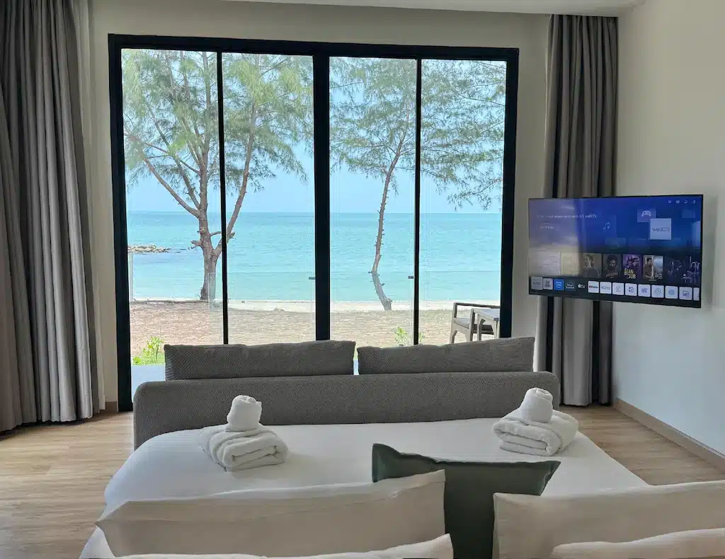 ห้องพักโรงแรม ขนอม ที่ทันสมัยพร้อมวิวทะเลผ่านประตูบานกระจกขนาดใหญ่ โซฟา ผ้าเช็ดตัวบนเตียง และทีวีติดผนังที่แสดงเมนูสตรีมมิ่ง มอบการพักผ่อนที่สมบูรณ์แบบสำหรับการเข้าพักของคุณ.