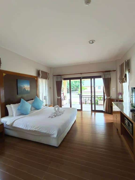 ห้องพักโรงแรมขนอมที่อบอุ่น พร้อมเตียงขนาดใหญ่ที่มีหมอนสีน้ำเงิน ตกแต่งด้วยผ้าขนหนูรูปหงส์ พื้นไม้ โทรทัศน์ โต๊ะทำงาน และประตูบานกระจกที่เปิดออกสู่ระเบียงพร้อมวิวทิวทัศน์ภายนอกที่สวยงาม.