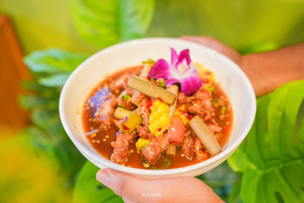 บุคคลหนึ่งถือชามแกงเผ็ดเพาะรักที่มีเนื้อสัตว์ ผัก ข้าวโพดอ่อน และตกแต่งด้วยดอกไม้สีม่วงที่รับประทานได้.