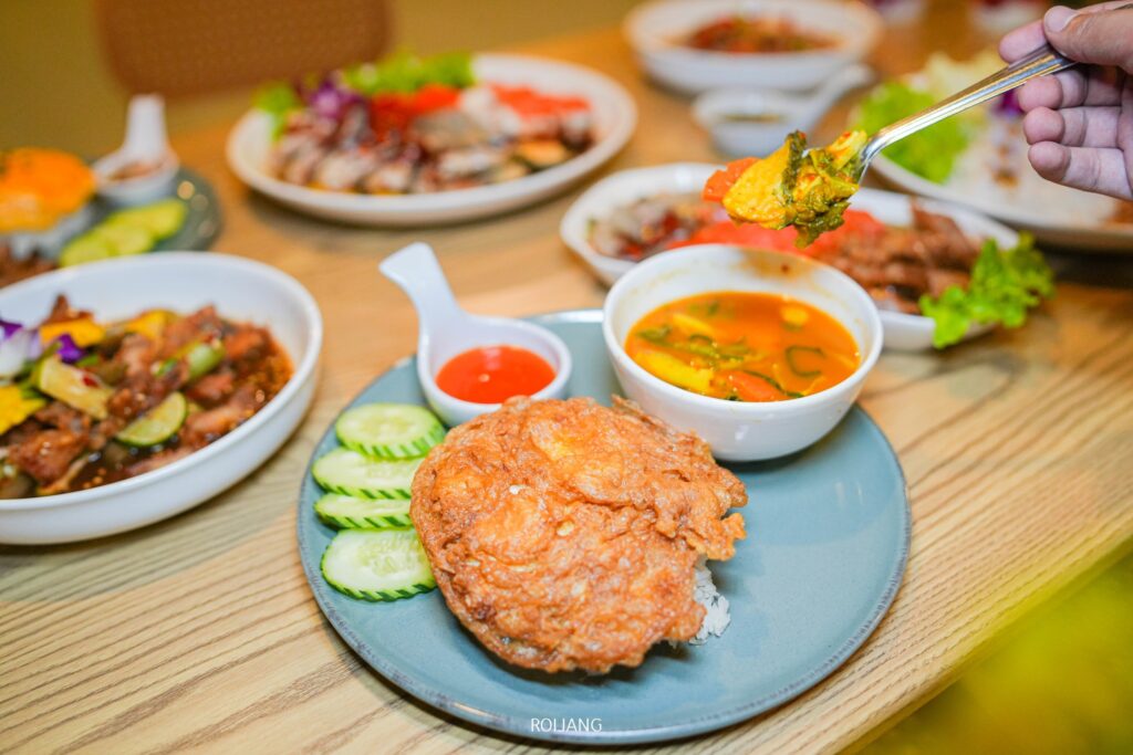 จานที่มีไข่เจียวทอด, แตงกวาหั่นเป็นแว่น, และถ้วยเล็ก ๆ ที่มีซอส, เสิร์ฟที่ เพาะรัก, พร้อมกับอาหารหลากหลายชนิด และมือที่กำลังถือส้อมที่มีอาหารอยู่ในพื้นหลัง.