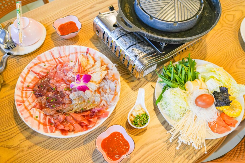 โต๊ะที่จัดเตรียมไว้พร้อมเตาแก๊สปิคนิค จานเนื้อและอาหารทะเลสดหลากหลายชนิด ถาดผักและเส้นบะหมี่ น้ำจิ้ม และเครื่องปรุงรส—เหมาะอย่างยิ่งสำหรับการรับประทานหม้อไฟในบรรยากาศอบอุ่นของ เพาะรัก.