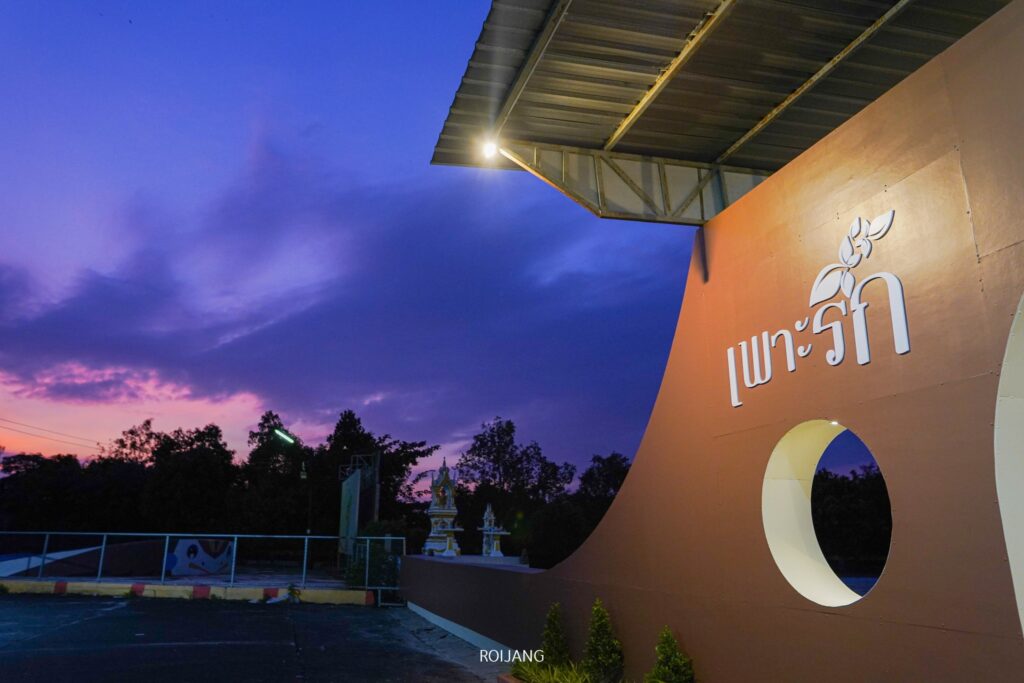 โครงสร้างโค้งที่มีข้อความภาษาไทย "เพาะรัก" พร้อมช่องตัดกลมและหลังคาปรากฏในภาพพระอาทิตย์ตกดิน ต้นไม้และท้องฟ้าสีม่วงอมน้ำเงินมองเห็นได้ในพื้นหลัง.