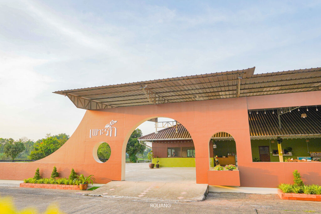 อาคารชั้นเดียวที่มีผนังโค้งสีส้ม ทางเข้าเปิดโล่ง และมีต้นไม้อยู่ด้านหน้า มองเห็นภายใต้ท้องฟ้าที่มีเมฆบางส่วน—เป็นพื้นที่ที่เชิญชวนซึ่งสะท้อนความอบอุ่นของ เพาะรัก ได้อย่างสมบูรณ์แบบ.