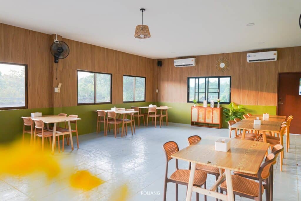 คาเฟ่สว่างไสวและโปร่งสบายชื่อว่า "เพาะรัก" มีโต๊ะและเก้าอี้ไม้ หน้าต่างบานใหญ่ ผนังสีเขียวและไม้ประดับ แอร์คอนดิชั่นติดตั้งอยู่ผนังด้านหลัง.