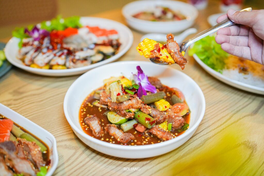 มือที่ถือช้อนซึ่งมีผักผสมและเนื้อย่างอยู่เต็มช้อน เหนือชามสลัดรสเผ็ดที่ร้าน เพาะรัก โดยมีอาหารหลากหลายชนิดจัดเรียงอยู่บนโต๊ะไม้.