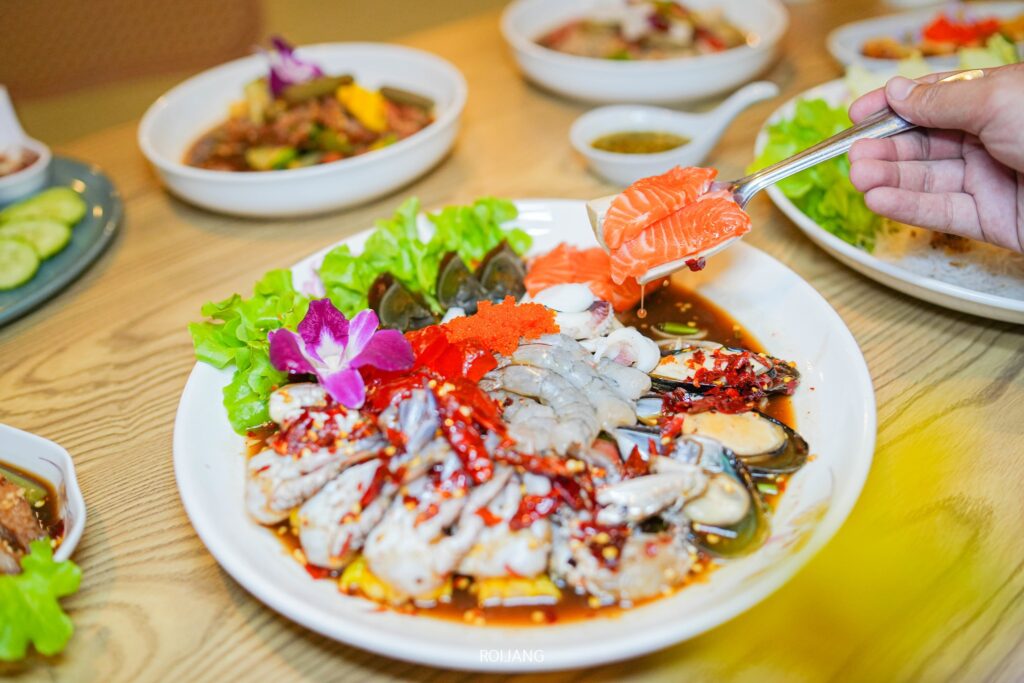 มือหนึ่งถือแซลมอนชิ้นหนึ่งไว้เหนือจานที่จัดเรียงอาหารทะเลสดหลากหลายชนิดจากร้าน เพาะรัก ประดับด้วยผักกาดหอม ดอกไม้ที่รับประทานได้ และซอสพริก บนโต๊ะไม้.