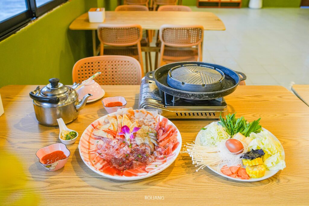 โต๊ะอาหารที่ร้าน เพาะรัก มีเตาปิ้งย่างหม้อไฟ, จานเนื้อและอาหารทะเลสด, ผักหลากหลายชนิด, กาต้มน้ำ, และชามซอสในบรรยากาศร้านอาหารที่อบอุ่น.