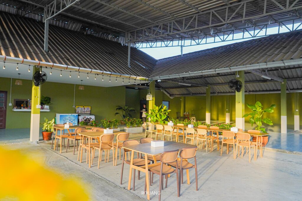 พื้นที่รับประทานอาหารกลางแจ้งที่ เพาะรัก พร้อมโต๊ะและเก้าอี้ไม้ใต้หลังคาเหล็ก ล้อมรอบด้วยผนังสีเขียว พืชพรรณเขียวชอุ่ม และไฟประดับแบบสายที่ดูมีเสน่ห์.