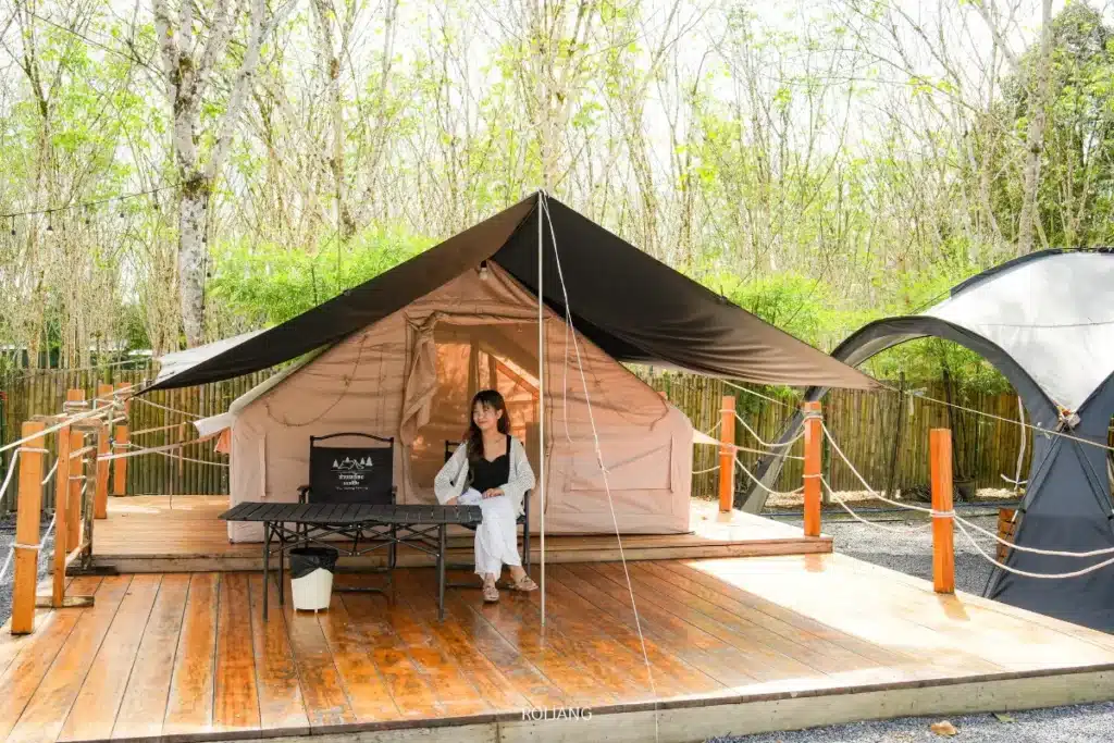 บุคคลหนึ่งนั่งอยู่กับมีสุขที่โต๊ะบนดาดฟ้าไม้หน้าเต็นท์ที่มีหลังคาลาดในบริเวณแคมป์ที่ล้อมรอบด้วยต้นไม้.