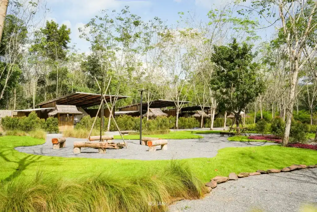 พื้นที่สวนสาธารณะกลางแจ้งที่มีม้านั่งไม้ ชิงช้า ทางเดินกรวด หญ้าเขียว และต้นไม้ที่กระจายอยู่ สร้างบรรยากาศที่ทุกคนรู้สึกมีความสุขท่ามกลางศาลาไม้ในฉากหลัง.