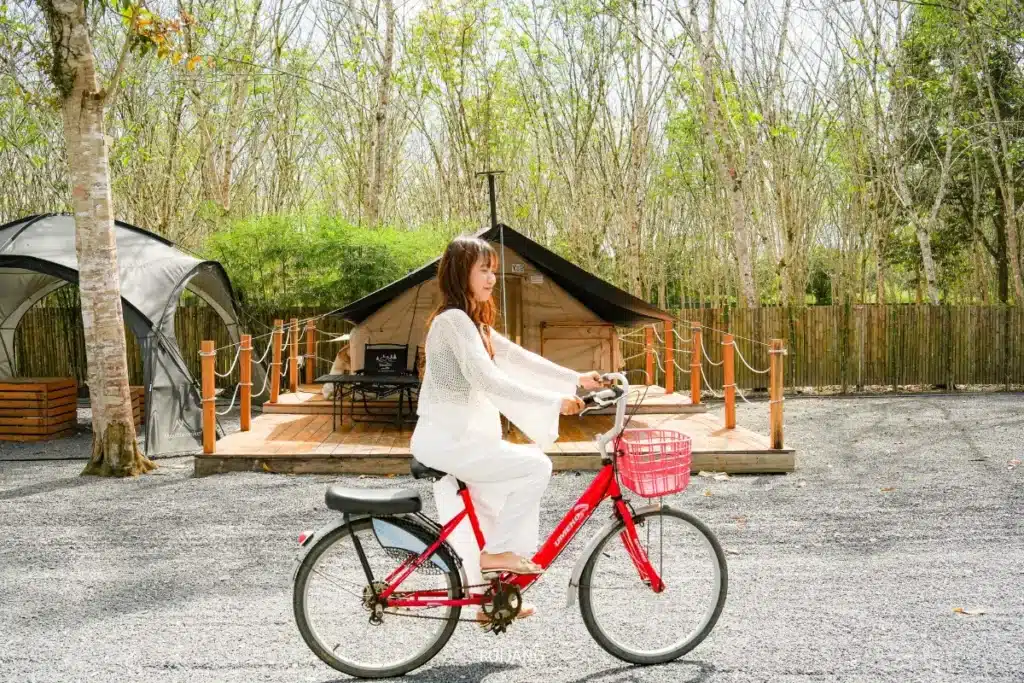 ผู้หญิงคนหนึ่งสวมเสื้อผ้าสีขาว ดูมีความสุข กำลังขี่จักรยานสีแดงผ่านกระท่อมไม้ในบรรยากาศกลางแจ้งที่เงียบสงบและมีต้นไม้ล้อมรอบ.