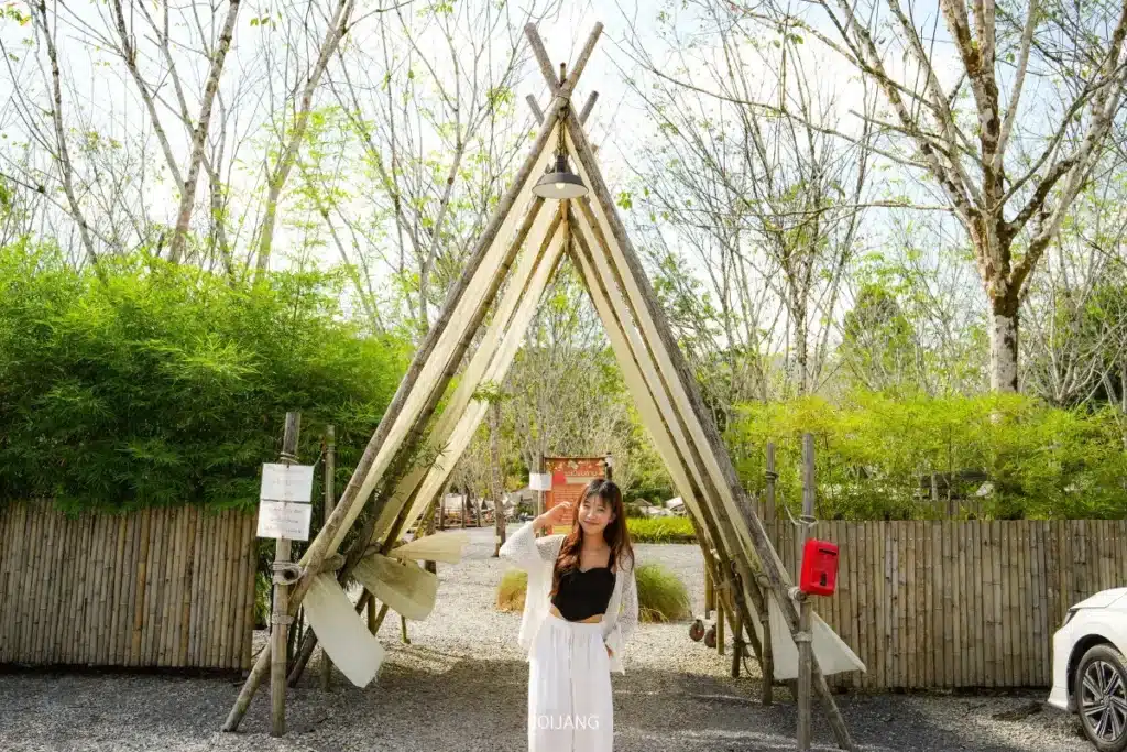 ผู้หญิงคนหนึ่งยืนอยู่ใต้โครงสร้างไม้รูปทรงเต็นท์ขนาดใหญ่กลางแจ้ง ล้อมรอบด้วยใบไม้สีเขียวและรั้วไม้ไผ่ในวันที่แดดจ้าและมีความสุข.