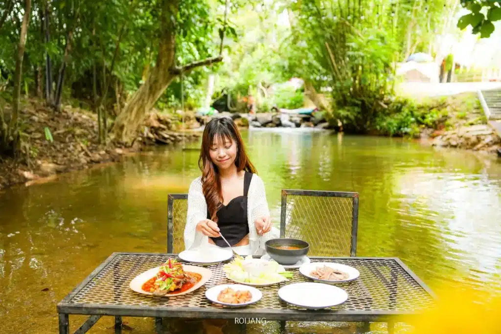 ผู้หญิงคนหนึ่งนั่งอยู่คนเดียวที่โต๊ะกลางแจ้งซึ่งจัดวางอาหารหลากหลายจานไว้ วางอยู่ในลำธารตื้นที่ล้อมรอบด้วยต้นไม้เขียวชอุ่ม กำลังเพลิดเพลินกับช่วงเวลาแห่งความสุขท่ามกลางธรรมชาติ.