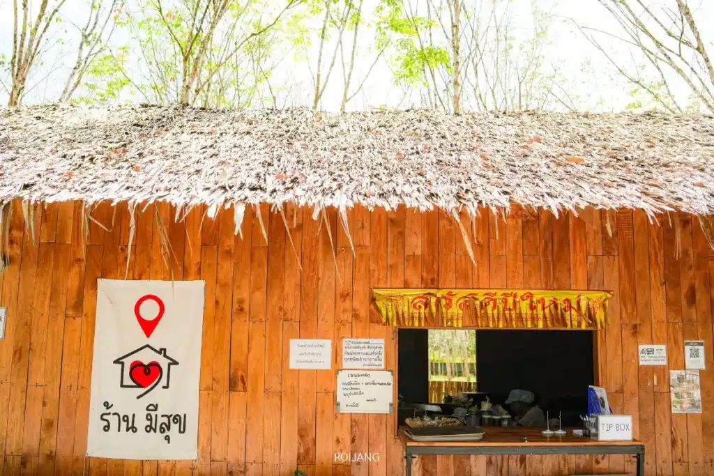 ร้านขายอาหารไม้หลังคามุงจาก มีป้ายเขียนด้วยอักษรไทยพร้อมคำว่า "มีสุข" อยู่ข้างสัญลักษณ์หมุดตำแหน่งที่ตั้ง และมีหน้าต่างบริการขนาดเล็กเปิดอยู่ซึ่งแสดงอาหารบางรายการ.