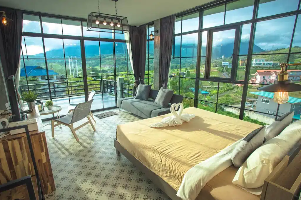 ห้องนอนสมัยใหม่ที่ โรงแรมเพชรบูรณ์ พร้อมหน้าต่างบานใหญ่ เตียงคู่ โซฟา และระเบียงที่มองเห็นภูเขาและตึกต่างๆ; แสงธรรมชาติส่องเข้ามาในห้อง.