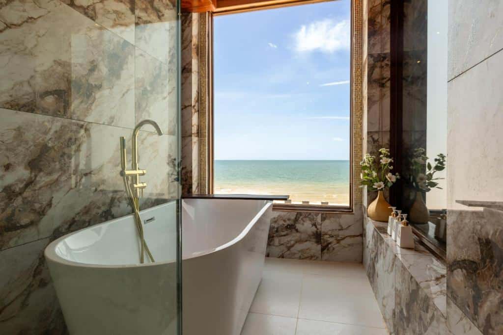 ห้องน้ำสไตล์โมเดิร์นพร้อมอ่างอาบน้ำแบบลอยตัวสีขาว อุปกรณ์ตกแต่งสีทอง ผนังหินอ่อน และหน้าต่างบานใหญ่ที่มองเห็นวิวทะเลและชายหาดทราย เหมาะอย่างยิ่งสำหรับการพักผ่อนหลังจากวันแห่งการสำรวจรีสอร์ทขนอม.