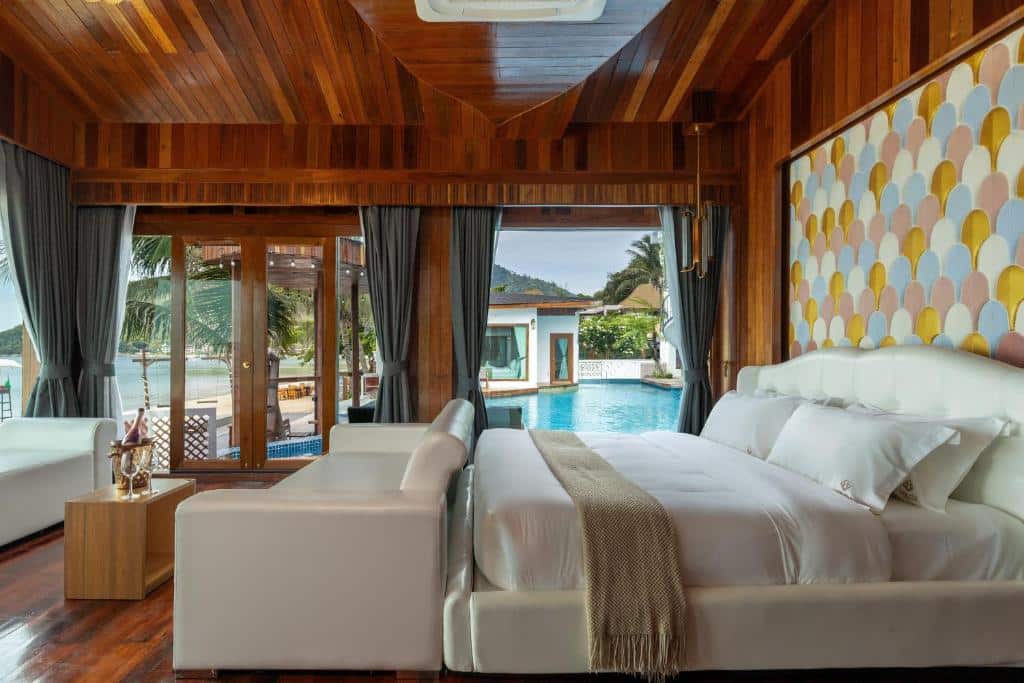 ห้องนอนสมัยใหม่ที่รีสอร์ทขนอม มีผนังและเพดานไม้ เตียงขนาดใหญ่ โซฟาสีขาว งานศิลปะเหนือเตียง และประตูบานกระจกที่เปิดออกสู่สระว่ายน้ำและพื้นที่กลางแจ้งที่เขียวชอุ่มด้วยพืชเขตร้อน.