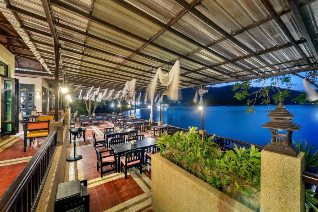 ระเบียงร้านอาหารกลางแจ้งที่มีหลังคาคลุม พร้อมโต๊ะและเก้าอี้ไม้ มองเห็นผืนน้ำที่สงบในยามเย็น—เหมาะสำหรับแขกที่กำลังมองหาที่พักในขนอมหรือค่ำคืนที่ผ่อนคลายในรีสอร์ทในขนอม พร้อมด้วยพืชพรรณและแสงไฟนวลตาที่ช่วยเสริมบรรยากาศอันเงียบสงบ.