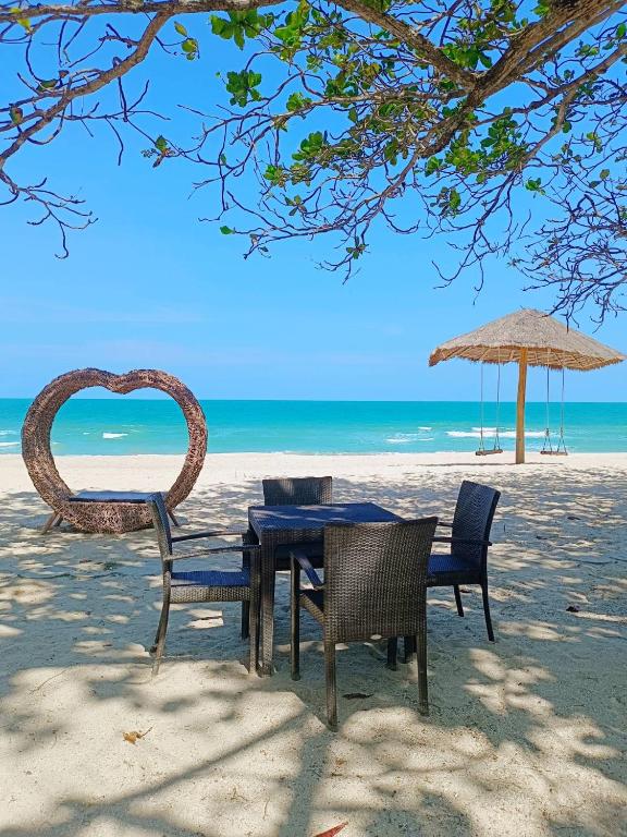 ชุดเก้าอี้หวายสี่ตัวและโต๊ะหนึ่งตัวบนชายหาดทรายที่รีสอร์ทขนอม พร้อมโครงหัวใจหวาย ชิงช้า และร่มจากหญ้าคาใกล้ทะเลภายใต้ท้องฟ้าสีฟ้าใส.