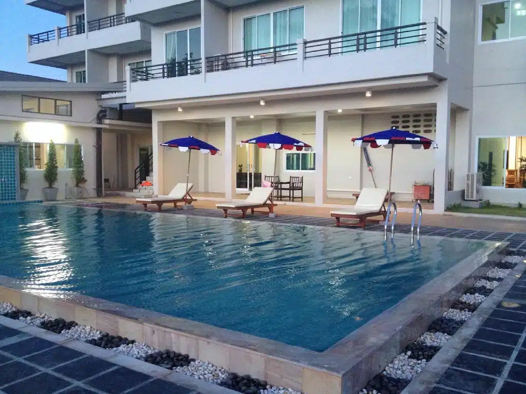 สระว่ายน้ำกลางแจ้งพร้อมเก้าอี้อาบแดดสี่ตัวใต้ร่มสีฟ้า ตั้งอยู่หน้าตึกสูงหลายชั้นสไตล์โมเดิร์นที่มีระเบียง—เหมาะสำหรับการพักผ่อนที่ ที่พักขนอม แห่งนี้.