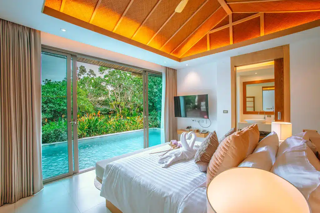 ห้องพักโรงแรมสมัยใหม่ที่ โรงแรมขนอม พร้อมเตียงที่จัดไว้อย่างเรียบร้อย การตกแต่งด้วยผ้าขนหนูรูปหงส์ โทรทัศน์ขนาดใหญ่ ประตูกระจกที่เปิดออกสู่สระว่ายน้ำส่วนตัว และต้นไม้สีเขียวด้านนอก.