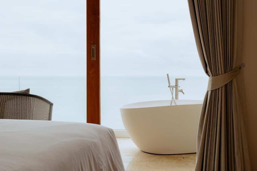 ห้องนอนสมัยใหม่ที่รีสอร์ทขนอมมีเตียงนอนที่แสนสบาย, เก้าอี้หวาย, และอ่างอาบน้ำแบบตั้งอิสระอยู่ข้างหน้าต่างกระจกบานใหญ่ที่มองเห็นทิวทัศน์ทะเลที่สงบและมีเมฆปกคลุม.
