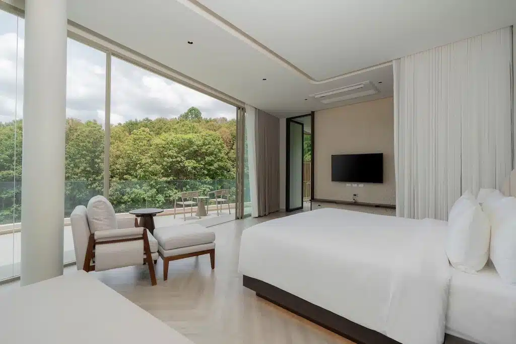 ห้องพักโรงแรมเพชรบูรณ์สมัยใหม่พร้อมเตียงขนาดใหญ่, เก้าอี้พักผ่อน, โทรทัศน์, และหน้าต่างบานใหญ่จากพื้นจรดเพดานที่สามารถมองเห็นระเบียงและต้นไม้สีเขียวภายนอกได้, เหมาะสำหรับการพักผ่อนอย่างผ่อนคลายในเพชรบูรณ์.