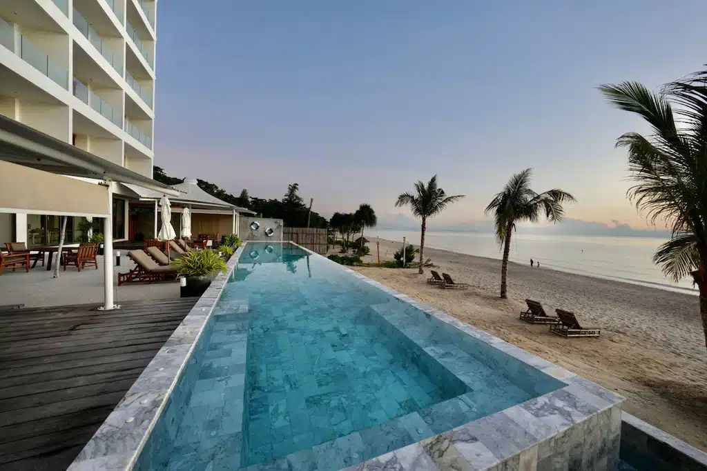 สระว่ายน้ำไร้ขอบพร้อมขอบหินอ่อนมองเห็นชายหาดทรายที่เรียงรายไปด้วยต้นปาล์ม; โรงแรมขนอม และเก้าอี้อาบแดดที่มองเห็นทางซ้ายมือในยามพระอาทิตย์ขึ้นหรือพระอาทิตย์ตก.