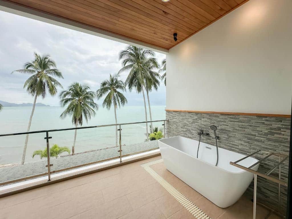 อ่างอาบน้ำสีขาวตั้งอยู่บนระเบียงที่มองเห็นวิวทะเลพร้อมต้นปาล์มที่ โรงแรมขนอม ซึ่งมีพื้นกระเบื้องและเพดานไม้.